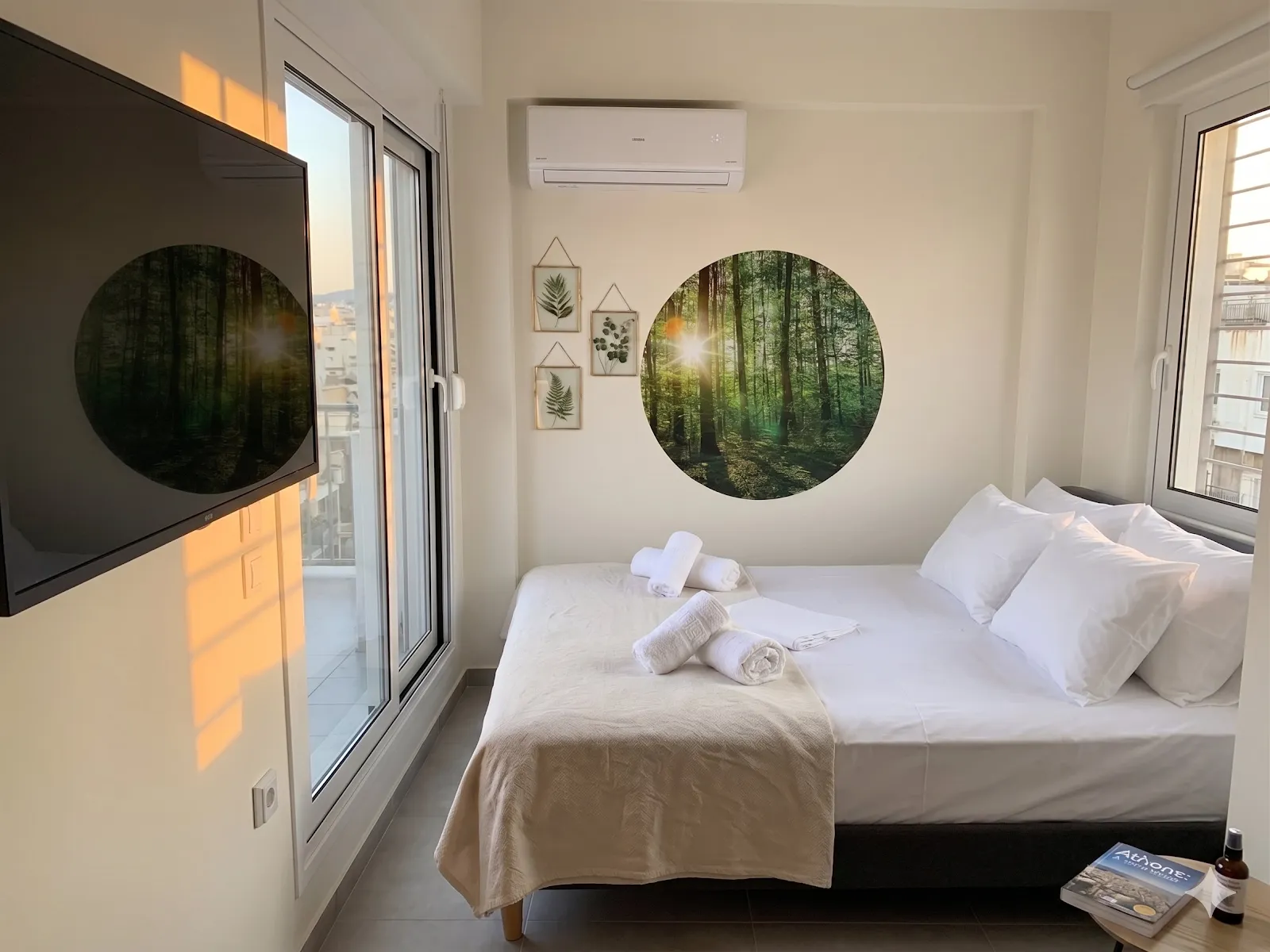 Bright bedroom with crisp white linen, a circular forest print above the bed, a wall-mounted television, and balcony doors open to the afternoon light.